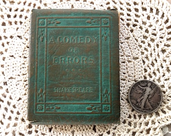 Pode incluir: Um pequeno livro retangular de cor verde-azulada intitulado A Comedy of Errors de Shakespeare, com detalhes florais em relevo na capa. Uma moeda de prata de meio dólar Walking Liberty de 1939 está posicionada à direita para escala.