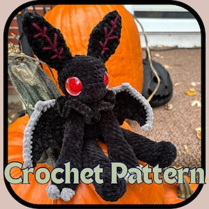 May include: A black crocheted plush toy shaped like a moth with large red eyes, pointed ears featuring dark red stitched details, and grey-tipped wings. The creature has a soft, fuzzy texture and is positioned in front of a large orange pumpkin.