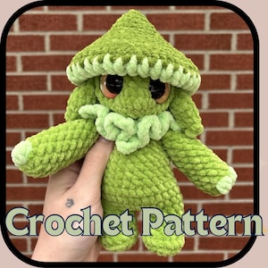 Puede incluir: Una criatura de ganchillo verde afelpado con un sombrero ancho y texturizado en forma de hongo, ojos grandes y oscuros y un cuello con volantes. La figura está hecha de hilo suave y difuso con detalles en verde claro. El texto dice Crochet Pattern.