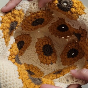 May include: A cream-colored crochet hat featuring a sunflower design. The sunflowers have yellow petals and brown centers. The hat is handmade with a textured stitch, showcasing the detailed craftsmanship. The hat is held by two hands.