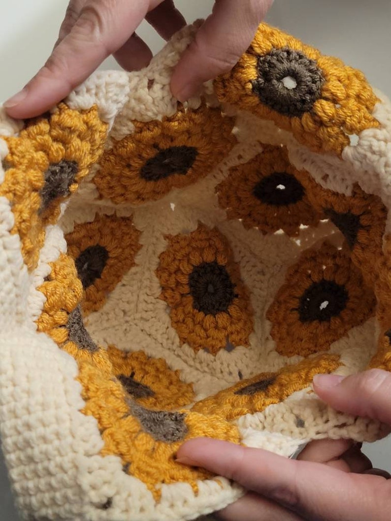 May include: A cream-colored crochet hat featuring a sunflower design. The sunflowers have yellow petals and brown centers. The hat is handmade with a textured stitch, showcasing the detailed craftsmanship. The hat is held by two hands.