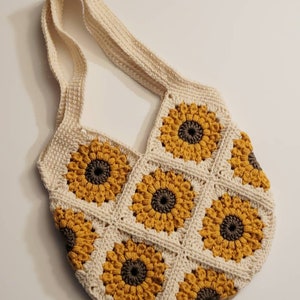 May include: Cream-colored crochet shoulder bag featuring a sunflower design. The bag is made of individual squares, each with a yellow sunflower and a brown center. The strap is also cream-colored.