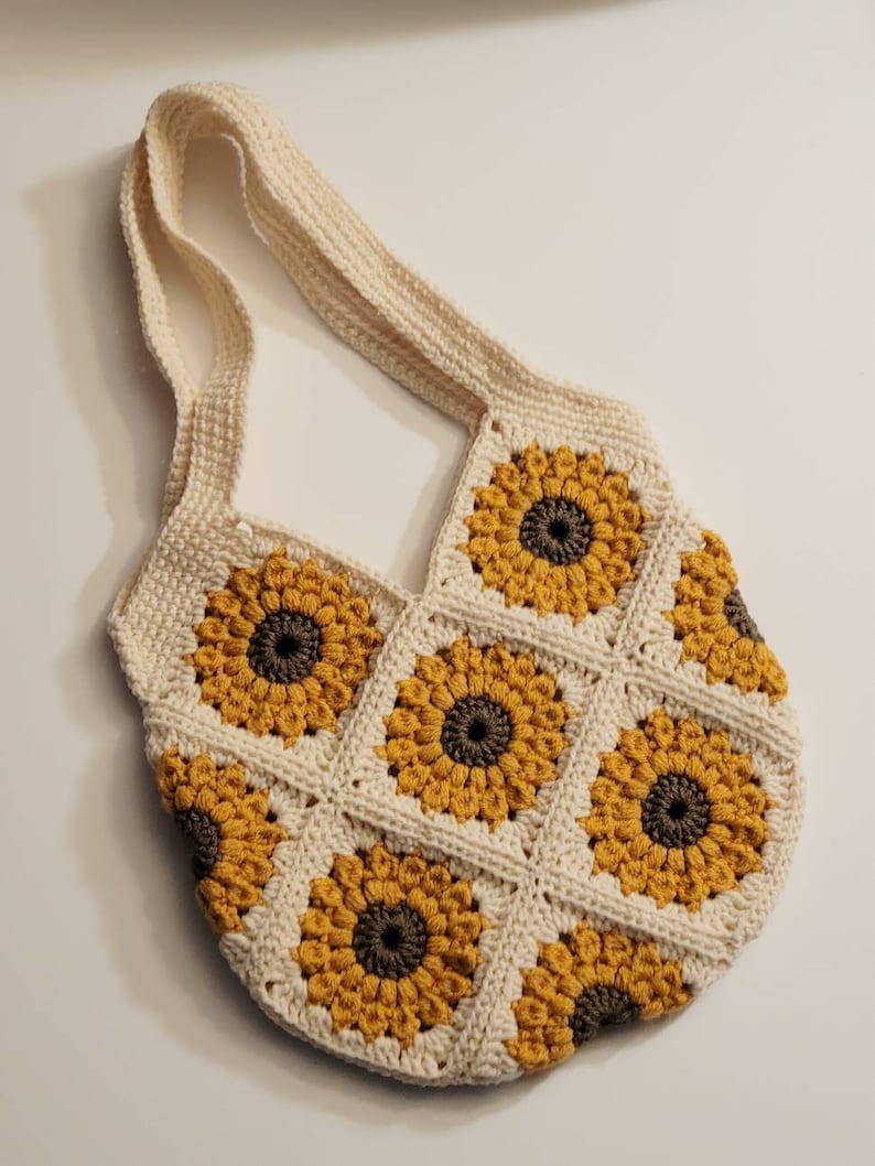 May include: Cream-colored crochet shoulder bag featuring a sunflower design. The bag is made of individual squares, each with a yellow sunflower and a brown center. The strap is also cream-colored.