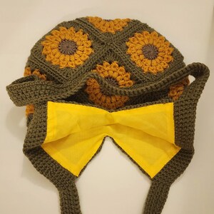 May include: A handmade crochet bag featuring a sunflower design. The bag is primarily olive green with yellow and brown sunflower motifs. The interior lining is bright yellow, and the bag has two straps.