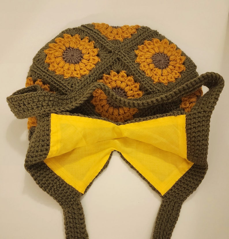 May include: A handmade crochet bag featuring a sunflower design. The bag is primarily olive green with yellow and brown sunflower motifs. The interior lining is bright yellow, and the bag has two straps.