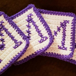 May include: Four handmade crochet coasters. Each coaster is square, featuring a white center with a purple letter "M" design. The edges are trimmed with a matching purple border. The coasters are arranged in a slightly overlapping pattern.