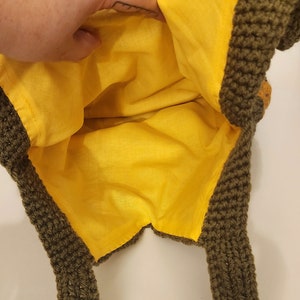 May include: A handmade crochet bag with a bright yellow interior lining. The exterior is a dark olive green color. The bag has two straps and a decorative element on the side. The bag is open, revealing the yellow lining.