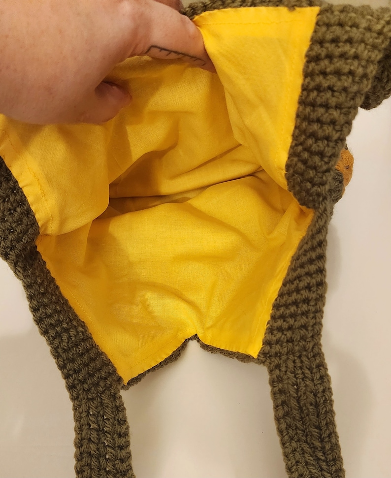 May include: A handmade crochet bag with a bright yellow interior lining. The exterior is a dark olive green color. The bag has two straps and a decorative element on the side. The bag is open, revealing the yellow lining.