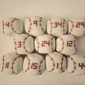 May include: A collection of eleven white crocheted baseballs with red stitching and numbers. The numbers, ranging from 3 to 44, are embroidered in red. The baseballs are arranged in a grid pattern against a plain background.
