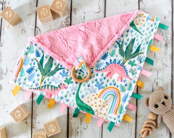 May include: A pink and white baby blanket with a dinosaur and rainbow print. The blanket has a soft, fuzzy texture and is trimmed with colorful ribbons. A wooden teether ring is attached to the blanket.