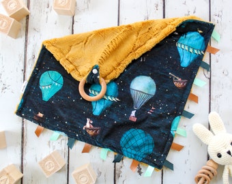 May include: A soft, plush baby blanket with a mustard yellow side and a dark blue side with a hot air balloon print. The blanket has a wooden ring attached to it and several colorful ribbons.