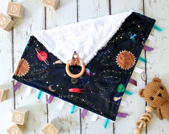 May include: A soft, black and white baby blanket with a space theme. The blanket features a sun, planets, stars, and a rocket. It has a white, fluffy side and a black side with a space print. The blanket has several colorful ribbons attached to the edges.