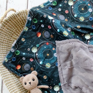 May include: A soft, plush baby blanket with a dark blue background featuring a celestial pattern of planets, stars, and swirling lines. The blanket is paired with a gray, fluffy minky fabric on the reverse side. A small, crocheted teddy bear is sitting on the blanket.