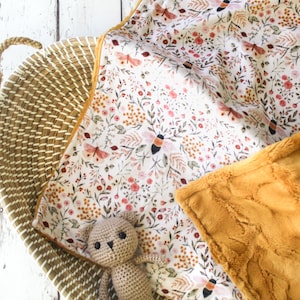 May include: A soft, plush baby blanket with a floral and bee print on one side and a solid mustard yellow faux fur on the other side. A small crocheted teddy bear is lying on the blanket.