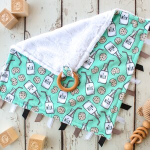 May include: A turquoise and white baby blanket with a milk bottle and cookie pattern. The blanket has several black and gray ribbons attached to the edges. A wooden teething ring is attached to the blanket.