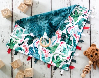 May include: A teal and white baby blanket with a dragon print and red and green ribbon ties. The blanket has a wooden teething ring attached.