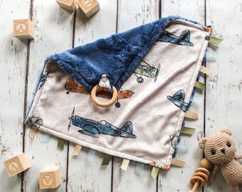 May include: A baby blanket with a blue plush border and a cream-colored center featuring airplane illustrations in shades of blue, orange, and green. The blanket has a wooden ring and multiple colored ribbons. Wooden blocks with letters are scattered around.