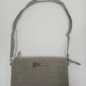 Puede incluir: Un bolso bandolera gris con una correa larga y fina. El bolso tiene forma rectangular con cierre de cremallera. Hay texto impreso en la parte delantera. La correa está sujeta con herrajes metálicos plateados.