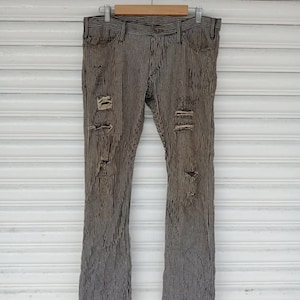 May include: Distressed, vertically striped denim jeans in shades of brown and white. The jeans have multiple rips and tears across the front of the legs. The pants are hanging from a wooden hanger.