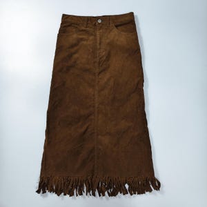 May include: A dark brown suede midi skirt featuring a high-waisted design with a button closure, a front zipper, two side pockets, and a long fringe hemline that extends from the bottom edge of the skirt, creating a textured and layered appearance.