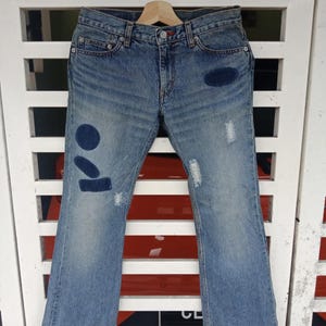 May include: Light blue denim jeans with a faded wash and distressed details. The jeans feature patches with a circle and oval design on the left leg. The jeans are hanging on a wooden hanger.