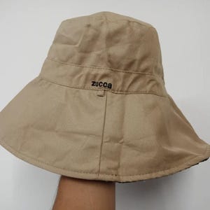 Puede incluir: Un sombrero de pescador beige con ala ancha. El sombrero tiene una corona estructurada y una banda en el medio. La palabra "zucca" está bordada en negro en la parte delantera. El sombrero está hecho de una tela resistente.