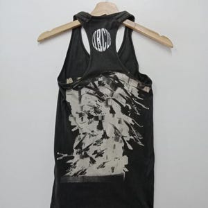 May include: Dark gray tank top with a white graphic design and the letters "RBC" on the back. The tank top is hanging on a wooden hanger against a white background. The graphic design appears to be abstract.