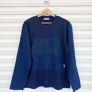 Puede incluir: Una camiseta de manga larga con cuello redondo en tonos azules. La parte superior es azul oscuro, con un parche azul claro similar al denim en el pecho. Las mangas son de material vaquero azul claro. La camiseta cuelga de una percha de madera.