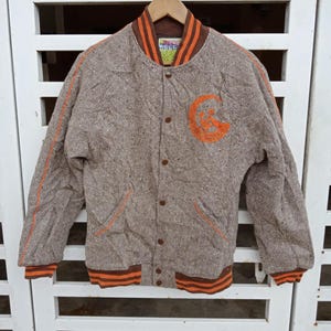 Puede incluir: Chaqueta universitaria marrón y naranja con un patrón de espiga. Cuenta con un parche naranja con la letra "G" en el pecho y rayas naranjas en las mangas y puños. La chaqueta tiene cierre de botones y cuello marrón.