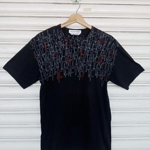 Puede incluir: Camiseta negra de manga corta con un diseño geométrico en blanco y rojo. El diseño cubre la parte superior de la camiseta. La camiseta está colgada de una percha de madera.