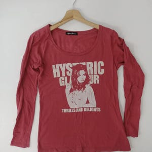 Puede incluir: Una camiseta roja de manga larga con cuello redondo. La camiseta presenta un gráfico de una mujer y las palabras "HYSTERIC GLAMOUR" y "THRILLS AND DELIGHTS" en blanco. Cuelga de una percha de madera.