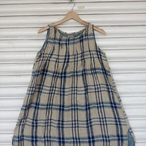 May include: A sleeveless, beige and navy plaid dress hangs on a wooden hanger. The dress features a loose, A-line silhouette with a split hem. The plaid pattern is composed of vertical and horizontal navy blue lines over a beige background.