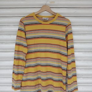 Puede incluir: Una camiseta amarilla de manga larga con rayas horizontales en varios colores, incluyendo rojo, gris y negro. La camiseta está colgada en una percha de madera sobre un fondo blanco. La camiseta tiene cuello redondo.