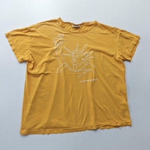 Puede incluir: Camiseta de color amarillo mostaza con un dibujo lineal blanco de la Estatua de la Libertad sosteniendo una antorcha. El texto "SPORTING GEAR HA!" está debajo. La camiseta tiene mangas cortas y cuello redondo.