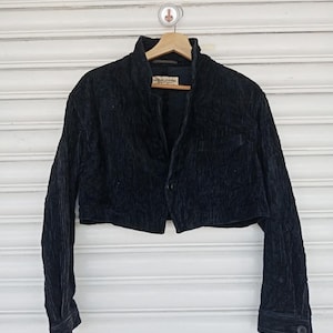 Puede incluir: Chaqueta de pana negra con diseño corto. La chaqueta tiene mangas largas, cierre de botones y cuello alto. Cuelga de una percha de madera sobre un fondo blanco.