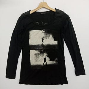 Puede incluir: Camiseta negra de manga larga con cuello redondo. La camiseta presenta una impresión fotográfica en blanco y negro de una persona caminando en un entorno brumoso. La camiseta está colgada de una percha de madera.