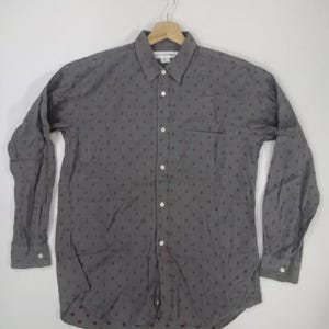 Puede incluir: Una camisa gris abotonada con un pequeño patrón de diamantes rojos repetidos. La camisa de manga larga tiene un bolsillo en el pecho y está colgada de una percha de madera. La camisa está abotonada y parece estar hecha de una tela ligera.