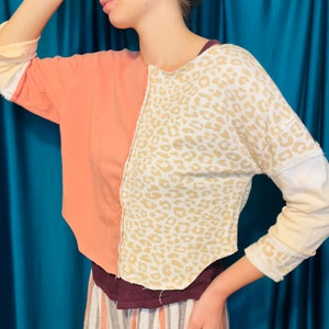 May include: A cropped, long-sleeved shirt with a peach-colored side and a white side with a brown leopard print. The shirt has a relaxed fit and a slightly asymmetrical hem.
