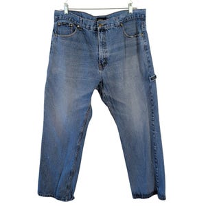 May include: Medium-wash blue denim jeans featuring a classic five-pocket design, a button and zipper fly closure, belt loops, and a utility loop on the left side. The jeans have a relaxed fit with subtle fading on the front thighs and legs.