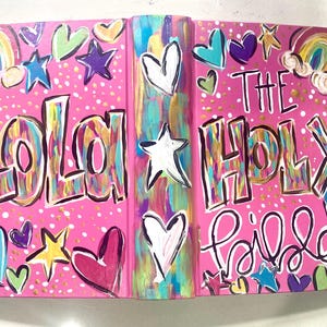 May include: A pink book cover with the words "LOLA" and "THE HOLY Bible" in a colorful, hand-painted style. The cover is decorated with hearts, stars, and rainbows.