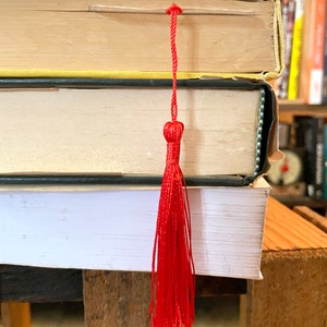 K&ouml;nnte beinhalten: Ein Stapel B&uuml;cher mit einem leuchtend roten Quasten-Lesezeichen. Die B&uuml;cher haben gelbe, schwarze und wei&szlig;e Einb&auml;nde. Das Lesezeichen besteht aus roten F&auml;den, die oben zusammengebunden sind und sich zu einer Quaste auff&auml;chern.