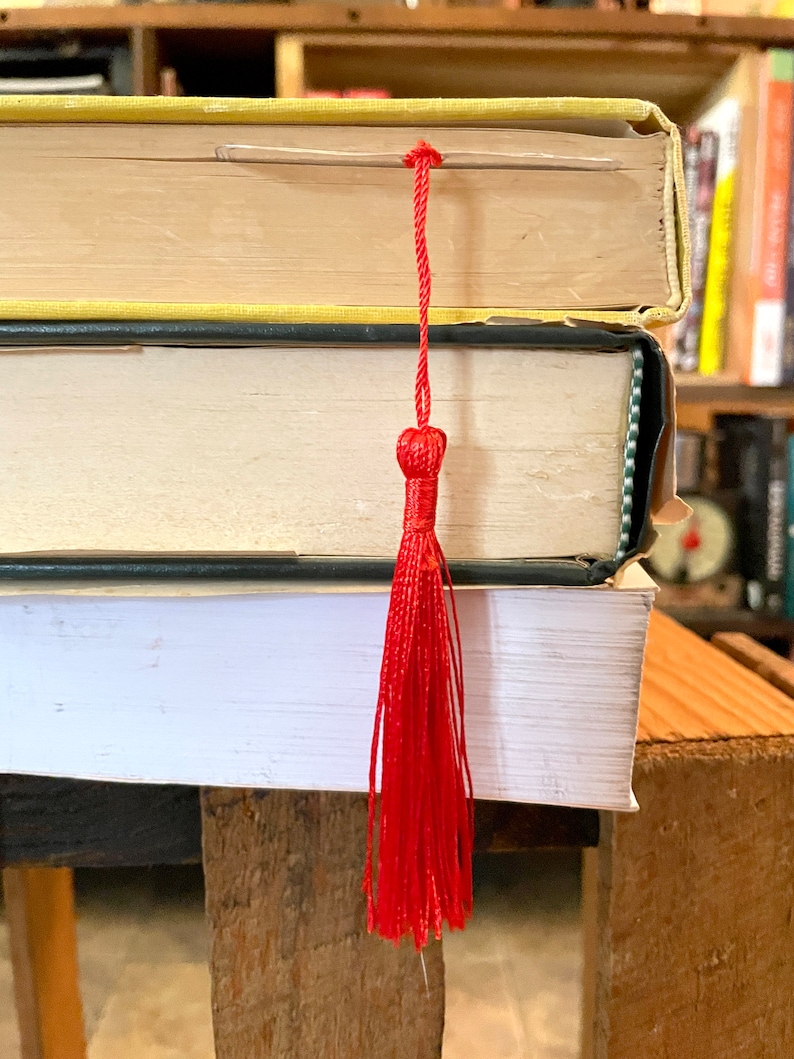 K&ouml;nnte beinhalten: Ein Stapel B&uuml;cher mit einem leuchtend roten Quasten-Lesezeichen. Die B&uuml;cher haben gelbe, schwarze und wei&szlig;e Einb&auml;nde. Das Lesezeichen besteht aus roten F&auml;den, die oben zusammengebunden sind und sich zu einer Quaste auff&auml;chern.