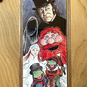 K&ouml;nnte beinhalten: Ein Lesezeichen mit einer farbenfrohen Illustration von Figuren aus einer Weihnachtsgeschichte. Die obere H&auml;lfte zeigt einen Mann mit Zylinder und eine Kapuzenfigur. Die untere H&auml;lfte zeigt zwei gr&uuml;ne froschartige Charaktere in Winterkleidung.