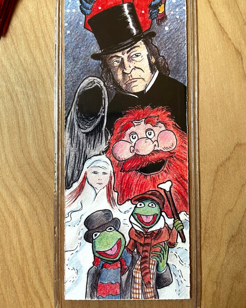 K&ouml;nnte beinhalten: Ein Lesezeichen mit einer farbenfrohen Illustration von Figuren aus einer Weihnachtsgeschichte. Die obere H&auml;lfte zeigt einen Mann mit Zylinder und eine Kapuzenfigur. Die untere H&auml;lfte zeigt zwei gr&uuml;ne froschartige Charaktere in Winterkleidung.