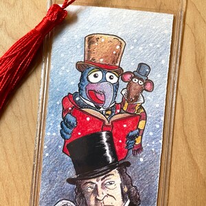 K&ouml;nnte beinhalten: Ein klarer Lesezeichen mit roter Quaste zeigt eine farbenfrohe Illustration von Gonzo und Rizzo die Ratte, gekleidet in viktorianischer Kleidung, die ein Buch lesen. Darunter befindet sich ein Portr&auml;t eines Mannes mit Zylinder. Das Lesezeichen ist in klarem Kunststoff eingeschlossen.