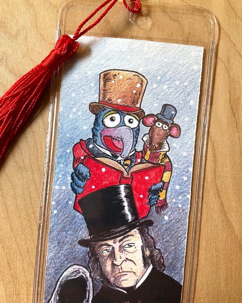 K&ouml;nnte beinhalten: Ein klarer Lesezeichen mit roter Quaste zeigt eine farbenfrohe Illustration von Gonzo und Rizzo die Ratte, gekleidet in viktorianischer Kleidung, die ein Buch lesen. Darunter befindet sich ein Portr&auml;t eines Mannes mit Zylinder. Das Lesezeichen ist in klarem Kunststoff eingeschlossen.