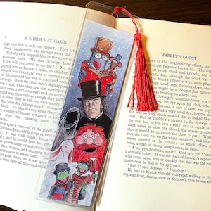 K&ouml;nnte beinhalten: Ein farbenfrohes Lesezeichen mit Illustrationen von Figuren aus "A Christmas Carol", darunter Scrooge und die Muppets. Es hat eine rote Quaste und steckt in einem Buch. Das Lesezeichen ist ca. 18 cm lang.