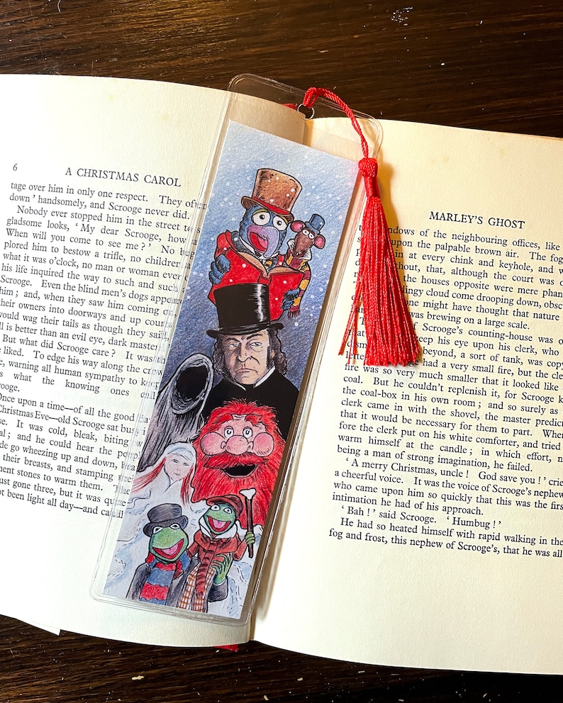 K&ouml;nnte beinhalten: Ein farbenfrohes Lesezeichen mit Illustrationen von Figuren aus "A Christmas Carol", darunter Scrooge und die Muppets. Es hat eine rote Quaste und steckt in einem Buch. Das Lesezeichen ist ca. 18 cm lang.