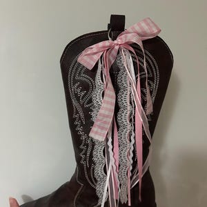 Op de afbeelding: Een bruine leren cowboylaars met witte stiksels en een decoratieve roze en wit geruite strik. De laars is versierd met roze, witte en kanten linten, wat een vrouwelijke touch geeft aan de schoenen in westerse stijl.