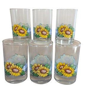 May include: Set of six clear glass tumblers. Each tumbler features a vibrant sunflower design with yellow petals, brown centers, and green leaves. The design is set against a blue and white checkered pattern. The glasses are ideal for serving beverages.